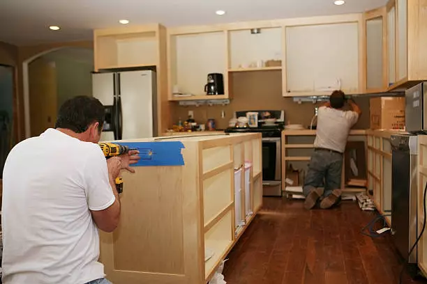 Cabinet maker installing custom made maple cabinets to kitchen island, while electrician installs under-cabinet lighting.